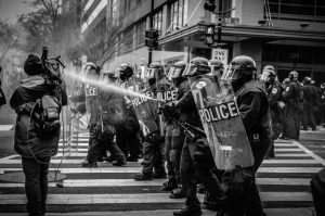 A black and white image of a line of policemen dressed in riot gear with shields and helmets stands in a city intersection and sprays water towards a cameraperson.